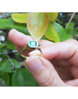 0.81 ct. emerald solitaire ring in 14K yellow gold, bezel set, held by fingers with green leaves.