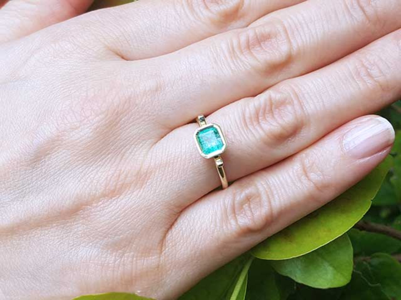 A solitaire emerald ring on a hand against a natural background