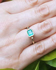 A solitaire emerald ring on a hand against a natural background