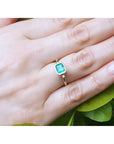 Emerald solitaire ring, bezel set in 14K yellow gold, worn on a hand with green leaves.
