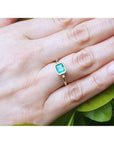 Emerald solitaire ring, bezel set in 14K yellow gold, worn on a hand with green leaves.