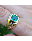 Gold ring for men with a square emerald-cut natural colombian emerald on a finger against a blurred background.