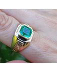 Gold ring for men with a square emerald-cut natural colombian emerald on a finger against a blurred background.