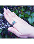 Hand wearing a ring for men with a natural emerald gemstone against a leafy background.