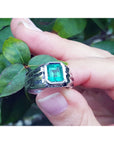 White gold ring for men with a Muzo colombian emerald held between fingers against a leafy background.