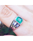 This white gold men's emerald ring is on a finger against a blurred natural background.