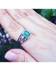 A white gold mens ring with a colombian emerald on a hand against a blurred natural background.