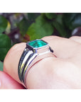 White gold mens ring with a genuine colombian emerald on a hand against a blurred green background.
