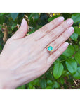 Hand wearing a gold emerald solitaire ring against a green leafy background.