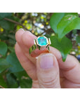 A Colombian emerald solitaire ring for women held by a hand against a green leafy background.