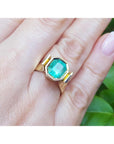 A women's emerald solitaire ring bezel set on a hand against a green leafy background.