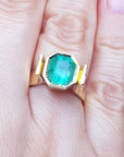 Emerald solitaire ring yellow gold on a hand, with green leaves in the background