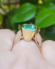 A bezel set emerald solitaire ring in yellow gold on a person's finger against a green leafy background.