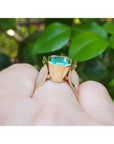 A bezel set emerald solitaire ring in yellow gold on a person's finger against a green leafy background.