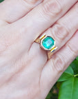 A 14k yellow gold emerald solitaire ring on a hand against a green leafy background.