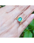 A 14k yellow gold emerald solitaire ring on a hand against a green leafy background.