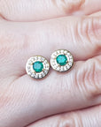 Gold earrings with emerald gemstones held between fingers on a light skin background.