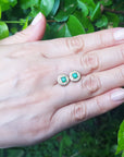 Hand holding a pair of emerald earrings with a green leafy background.