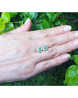 14k yellow gold emerald halo diamond stud earrings on a hand, deep bluish-green round emeralds.