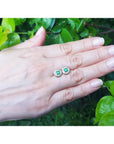 14k yellow gold emerald halo diamond stud earrings on a hand, deep bluish-green round emeralds.