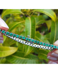 Natural emerald tennis bracelet held by a hand between fingers with a green leafy background.
