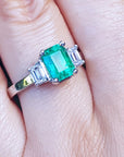 Emerald ring with emerald-cit side diamonds on a finger.