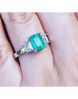 Emerald ring with emerald-cit side diamonds on a finger.