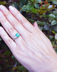 Hand wearing a natural emerald and side diamonds ring for women against a leafy background.
