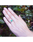 Three stone emerald ring with side diamonds in 18K white gold on a hand against green foliage.