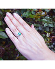 Three stone emerald ring with side diamonds in 18K white gold on a hand against green foliage.