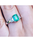 18K white gold three-stone emerald ring with a central bluish-green emerald and side emerald-cut diamonds.