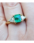 A yellow gold natural colombian emerald ring with side genuine trillion cut diamonds on a finger.