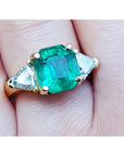 GIA Natural colombian emerald ring with trillion side diamonds in 18k yellow gold setting on a finger.