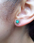 Close-up of a woman's ear wearing a Colombian emerald stud earring with a blurred background.