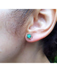 Close-up of a woman's ear wearing a Colombian emerald stud earring with a blurred background.