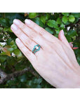 Hand wearing an enhancer emerald ring with a center diamond against a green leafy background.