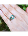 Hand wearing a ring with a pear-shaped center diamond and round emeralds gemstones against a blurred green background.