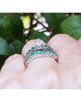 A white gold enhancer ring with emerald stones on a hand against a green leafy background.