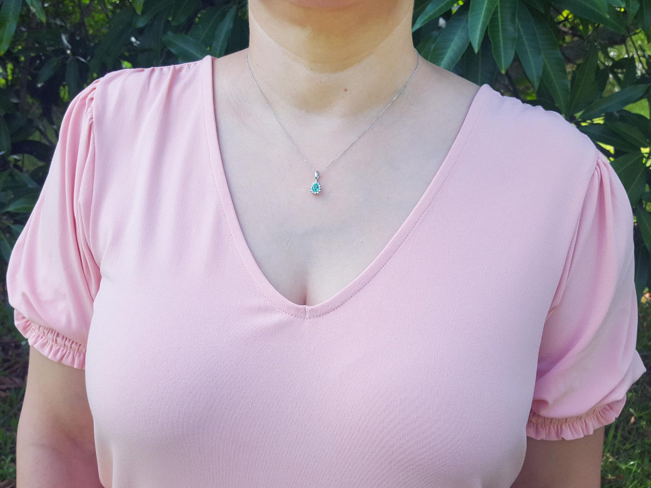 A woman wearing a light pink blouse with ruffled sleeves and an emerald enhancer pendant against a green leafy background.