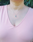 A woman wearing a light pink blouse with ruffled sleeves and an emerald enhancer pendant against a green leafy background.