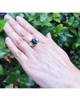 Hand wearing a gold ring with a black opal against a green leafy background.