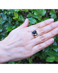 Hand wearing a ring with a black opal against a green leafy background.