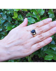 Hand wearing a ring with a black opal against a green leafy background.
