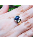 Gold ring with a black opal on a hand against a blurred green background.