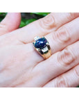 Gold ring with a black opal on a hand against a blurred green background.