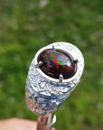 Men's silver ring with a colorful black Ethiopian black opal held against a blurred green background