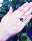 Hand wearing a sterling silver ring with a black Ethiopian black opal against a green leafy background.