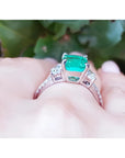 18K white gold ring with a cushion cut emerald and trapezoid side diamonds on a hand.