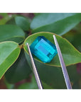 3.98 ct emerald-cut Muzo emerald, vibrant bluish-green, held by tweezers on a green leaf.