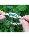 Gold bracelet with a Muzo colombian emerald held by a hand against a green leafy background